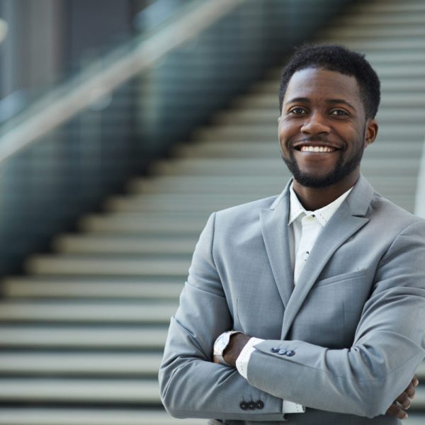 Portrait of jolly successful young black business executive with beard standing against staircase of lobby and crossing arms on chest